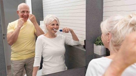 Illustrasjonsfoto: Eldre par står foran speilet på badet. Mannen bruker tanntråd og damen pusser tenner.