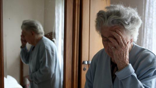 Fotoet viser en eldre dame i morgenkåpe som står på soverommet sitt. Hun tar seg på ansiktet med den venstre hånden og virker fortvilet.