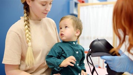 Fotoet viser en ung dame og en gutt (ca. 3 år), som sitter på fanget til kvinnen. Ha virker noe bekymre. På den høyre siden av fotoet vises det en del av hodet til en kvinnelig undersøker med rødt hår som holder en type VR-brille i sin venstre hånd. 