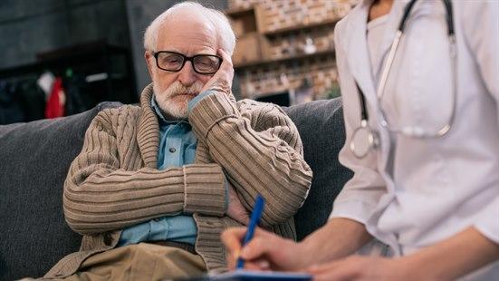 Illustrasjonsfoto av en eldre mann med trist uttrykk sitter i en sofa. Ved siden av sitter en person med hvit frakk og stetoskop og skriver.