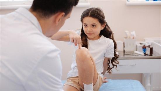 Illustrasjonsfotoet viser ei jente som er hos legen. Hun peker på kneet mens hun sitter på undersøkelsesbenken