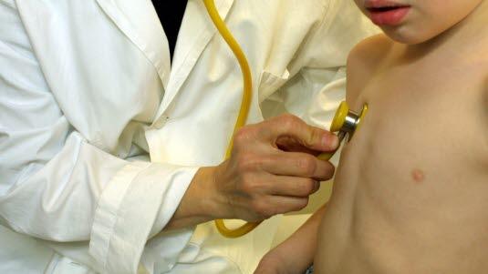 Fotoet viser en lege i hvit frakk som undersøker lungene til et yngre skolebarn med stetoskop. Bildeutsnittet viser overkroppene til de to personene. Barnet er i bar overkropp.