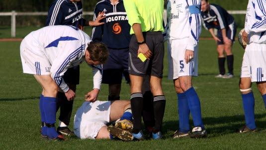 En fotballspiller har fått noe ut av ledd og ligger på banen, mens dommer og medspillere står rundt.