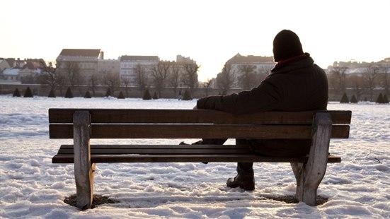 Illustrasjonsfoto viser omrisset av en person som sitter på en benk ute og ser mot en samling bygninger. Det er vinter.